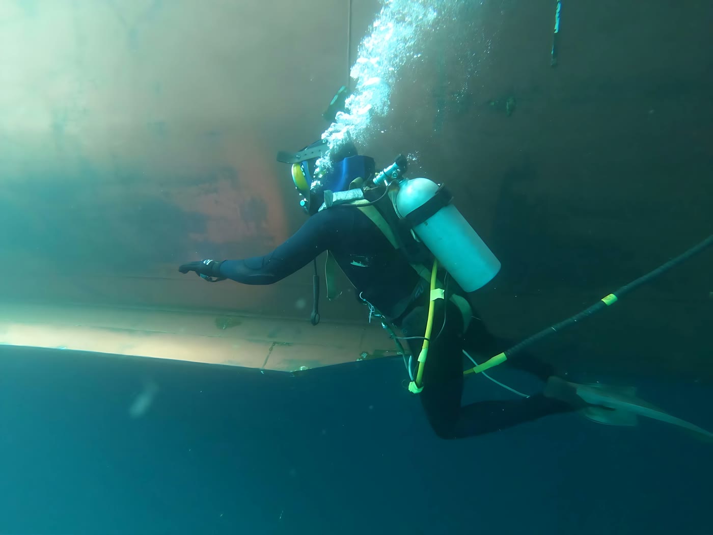Commercial diver inspecting a vessel hull underwater, trailing bubbles from their regulator