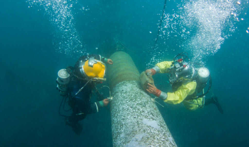 Two commercial divers inspecting and cleaning a subsea pipeline